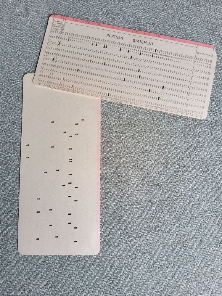 A couple of ancient computer punched cards, one face down showning blank side, the other face up showing "FORTRAN STATEMENT" heading and numbered column/row IDs. And a few holes punched in them.