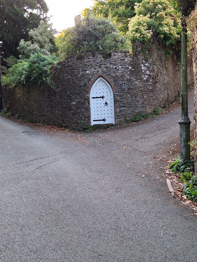 A white gate with a pointy top and solid but elegant black hinges and studs, set in the rounded end of a crenellated wall, where two lanes meet. Masses of greenery are on the other side of the wall.