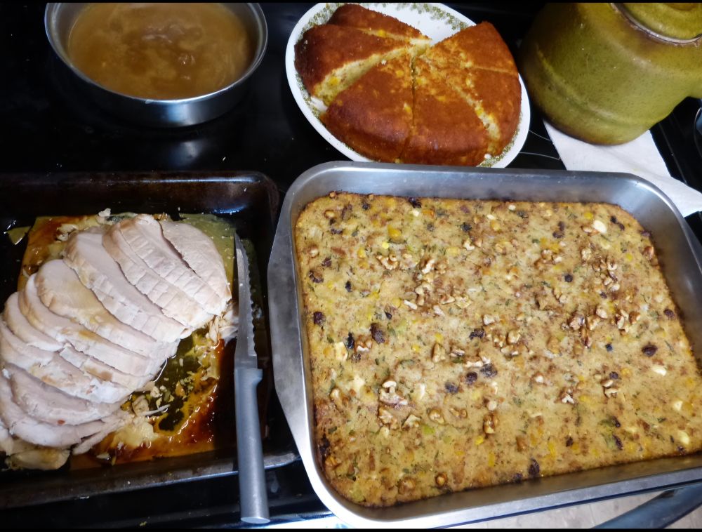 A stovetop with pans of cornbread dressing, sliced turkey and gravy. 