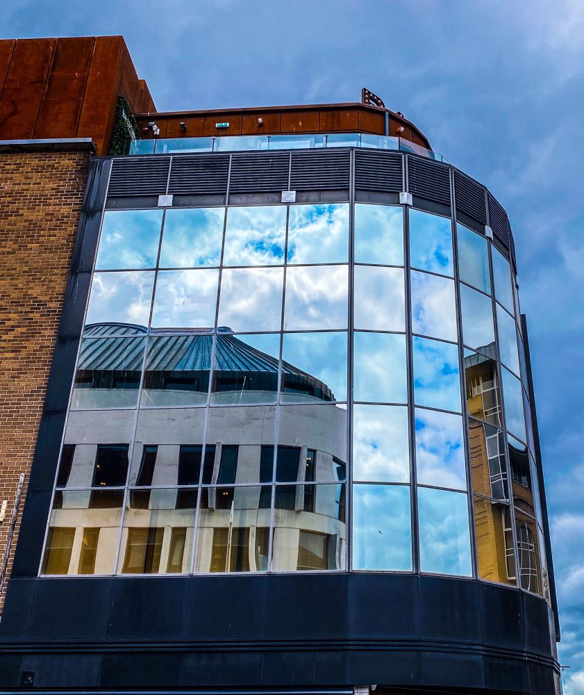 Looking up a building with glass panels on upper level walls. Blue sky and buildings opposite reflected in the glass