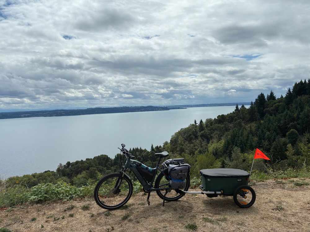 Ebike perched on top of a cliff pulling a green trailer full of camping supplies overlooking the Puget Sound facing south towards Tacoma