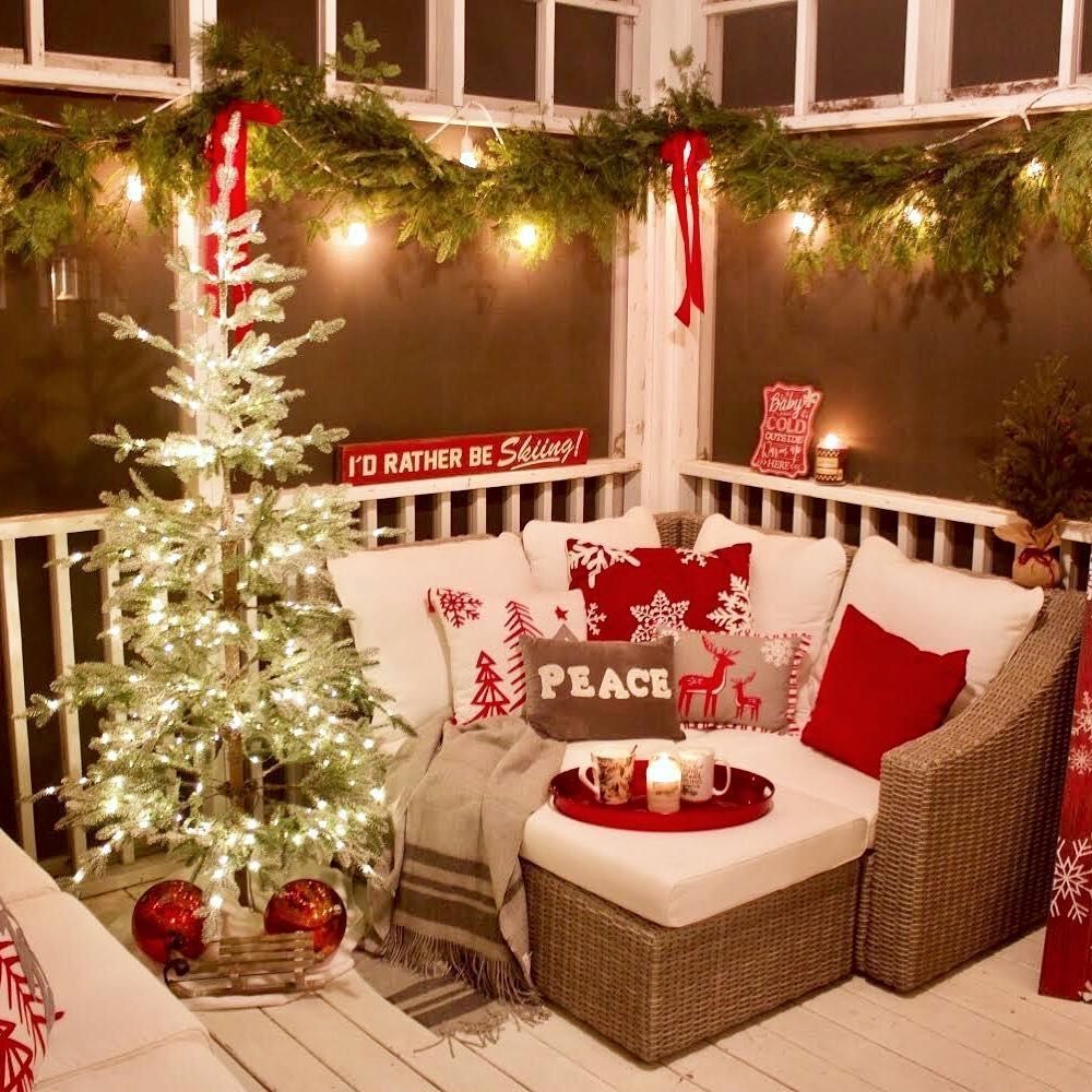 A small screened apartment porch fully decked out for Christmas with a 4-ft lit evergreen tree on the left, a corner wicker sofa with white and red Christmas cushions and evergreen and red bow lighted garland all around the top.