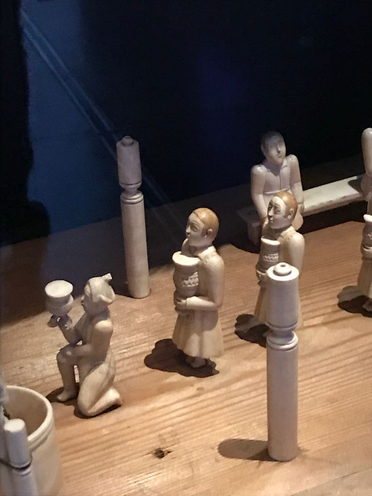 Several ivory figures on a wooden table. The one in front is kneeling and making an offering of a jar to a semi circular building. The two behind are standing and holding a jar. There are pillars either side of them. 