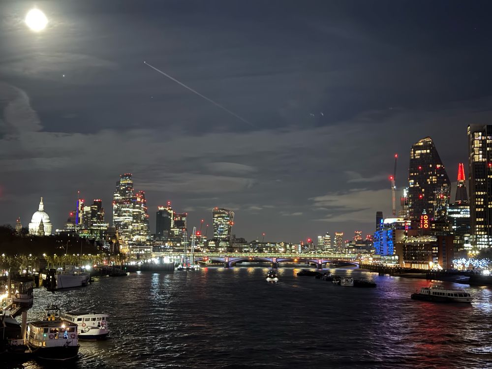 London at night. The river is in front, flowing away from us and the lights are reflected in the water. At the far right is St Paul’s, the white dome lit up. The moon is above it. In the distance are skyscrapers, lit up with gold and yellow and blue and red lights in the window. There is a shooting star in the clouds. 
