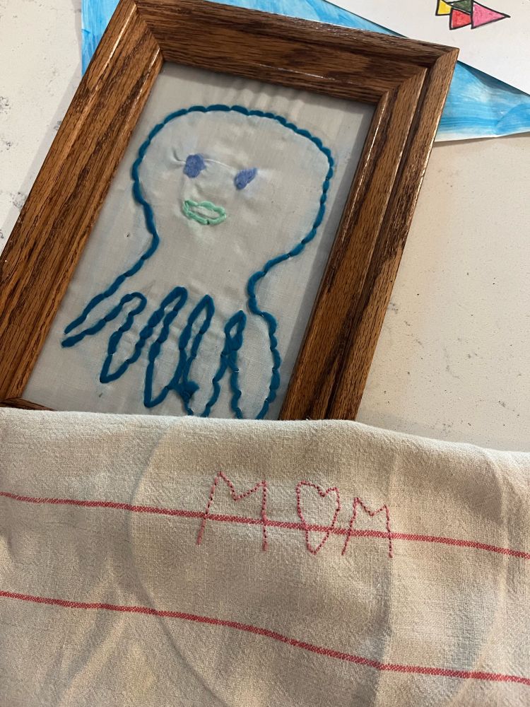 A blue octopus stitched with yarn in a frame and a tea towel stitched with the word MOM in pink thread. The O is a heart shape.