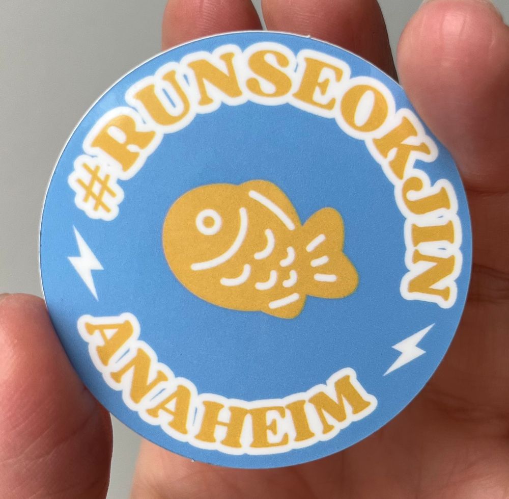 round sticker. sky blue background with yellow letters spelling #RUNSEOKJIN ANAHEIM and a bungeoppang in the center