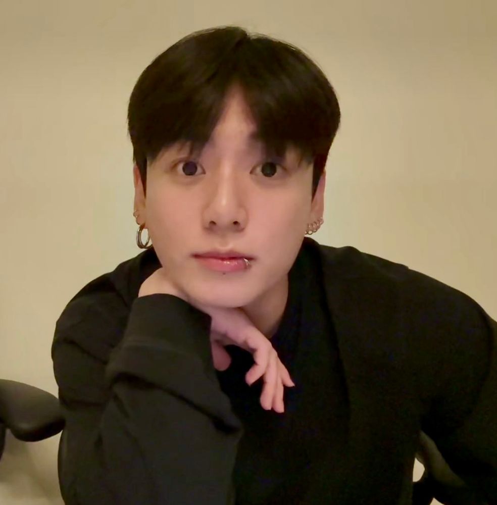 jungkook wearing a long sleeve black tee. he has his chin propped on a bent hand. his eyes are large and boba. 