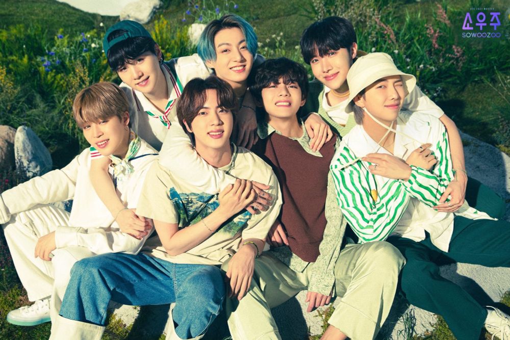 OT7 vegetable fritter sonyeondan 4ever. the seven members of BTS wearing casual outfits in shades of white, brown, green and denim, sitting in a grass field. the members are piled together in a group hug with warm smiles on their faces while the sun shines overhead.