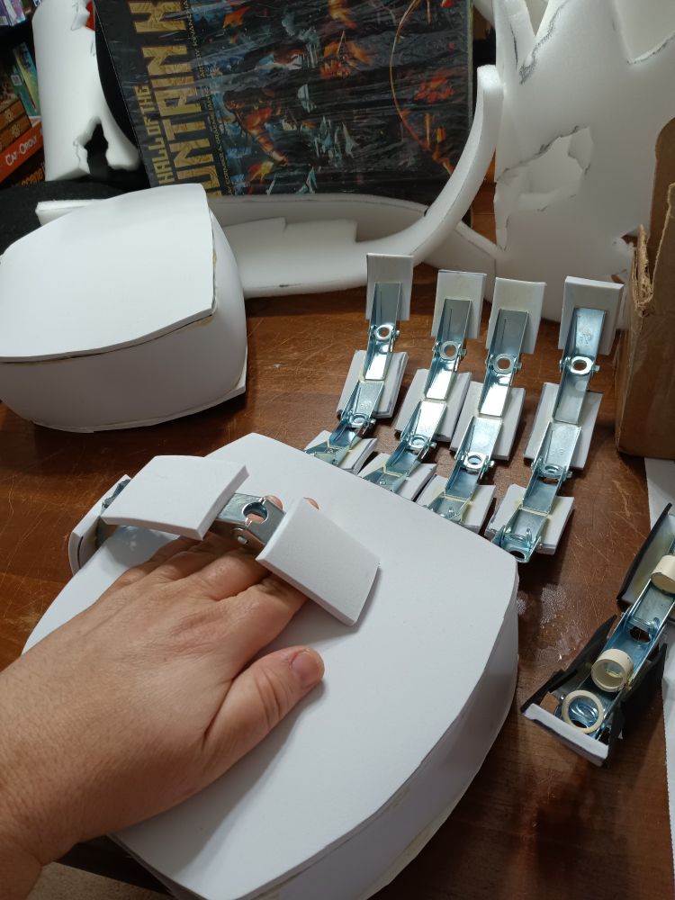 A hand for a cosplay suit. It is made of white foam, with silver metal bits making up the knuckle joints. It is resting palm up with a human hand on top of it for scale. The human hand is roughly one third the size of the cosplay one.
