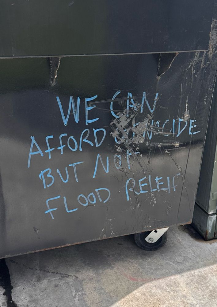 Graffiti on side of dumpster in Provo, Utah criticizing spending of public monies in the United States, "We can afford genocide but not flood relief".