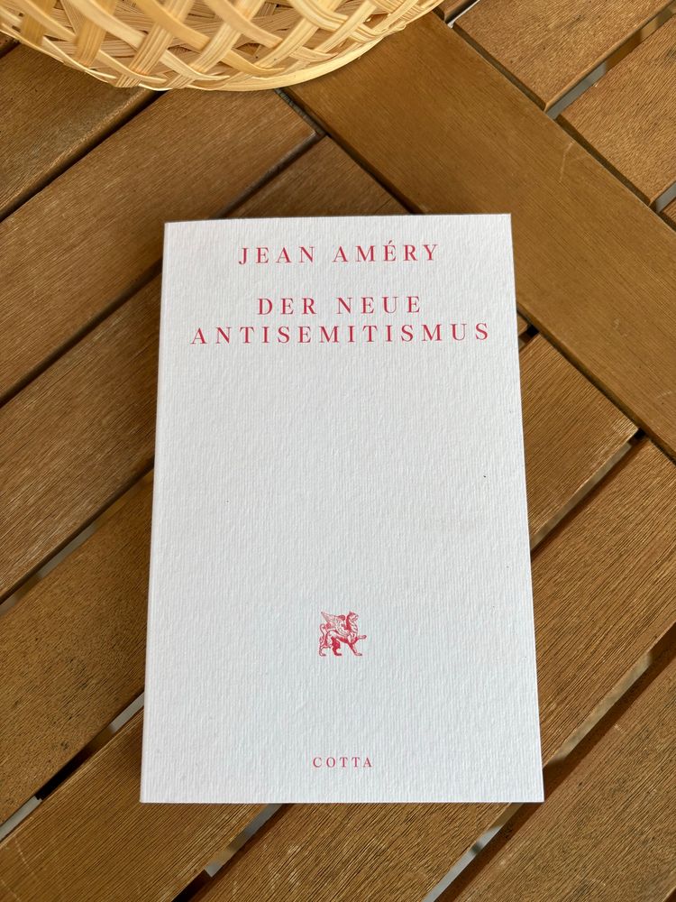 Ein Buch mit weißem Einband liegt auf einem hölzernen Gartentisch. Der Titel des Buches lautet „Jean Améry – Der neue Antisemitismus“, in roter Schrift zentriert auf dem Cover. Unten ist ein rotes Verlagslogo (ein geflügelter Löwe) und der Name des Verlags „COTTA“ zu sehen. Links oben im Bildrand ist ein Teil eines geflochtenen Korbes sichtbar.