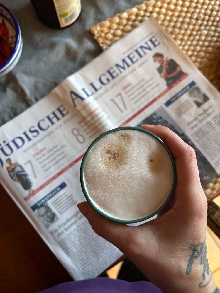 Eine Hand hält eine Tasse mit schaumigem Kaffee über einer auf dem Tisch liegenden Ausgabe der Zeitung „Jüdische Allgemeine“. Neben der Zeitung liegen ein geflochtener Untersetzer und teilweise sichtbare Gegenstände wie eine bunte Schale und eine Flasche. Die Hand hat eine Tätowierung am Handgelenk.