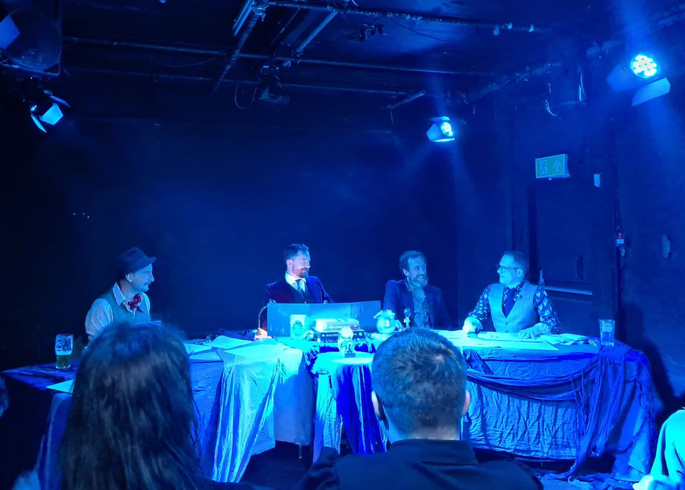 Four people on stage performing an actual play rpg. 3 players and a keeper sat in the centre.