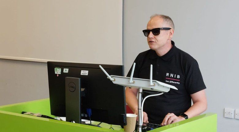 Dave Williams, RNIB’s Inclusive Design Ambassador, wearing an RNIB T-shirt presenting at a podium with a laptop and a microphone in a classroom setting