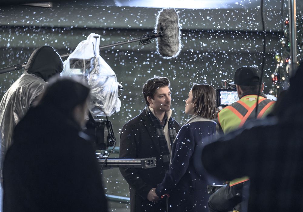 Behind the scenes, photo of the film set for Santa tell me, with Daniel Lissing and Erin Krakow in the center, surrounded by the film camera, monitors, and boom mic… as the snowfalls down around everyone