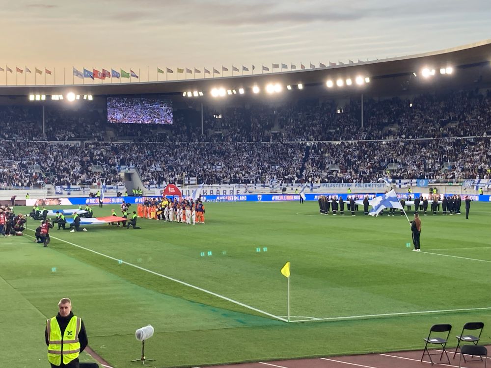 Beide teams staan vlak voor de wedstrijd op het veld naast elkaar. Linksonder staat een security meneer terwijl rechts iemand met een Finse vlag staat te wapperen.