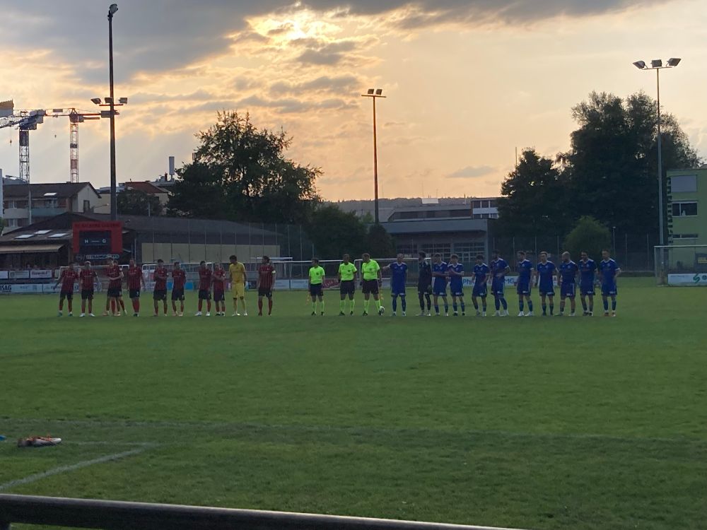 De line-up. FC Kickers speelt in het rood/zwart en FC Brunnen in het blauw. De scheidsrechters dragen felle gele kleuren. Op de achtergrond staan gebouwen.