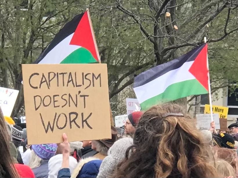 In a crowd, a protest sign has the text "Capitalism doesn't work," and there are two Palestinian flags waving in the wind.