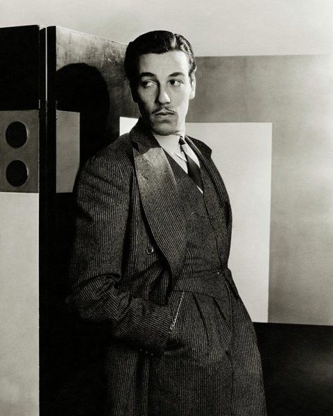 A young Cesar Romero in a three piece suit, hands in his pockets, leaning against a wall. His hair is slicked back and he has a small mustache