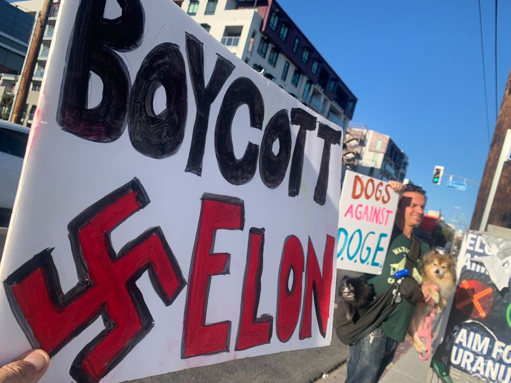 Two placards outside of protest at Elon Musk’s newly opened diner in West Hollwood. First placard reads “Boycott (swastika symbol) Elon.” Second placard held by man holding pet dog with placard that reads, “Dogs Against DOGE.”