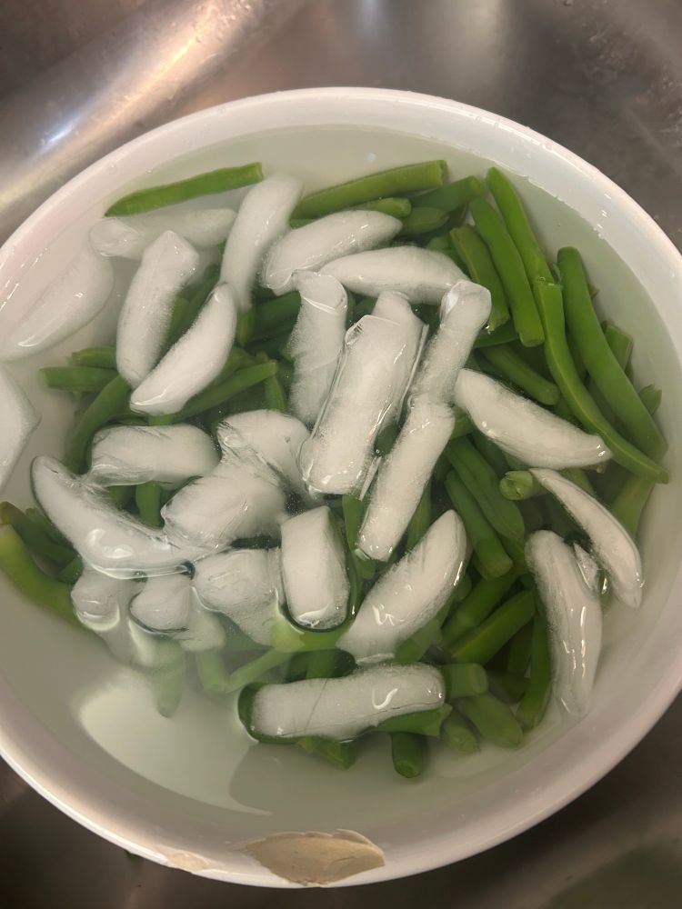 A white ceramic bowl with water and ice cubes floating around recently boiled green beans. A process called blanching, which keeps the color of the green beans from turning a bland olive shade. 