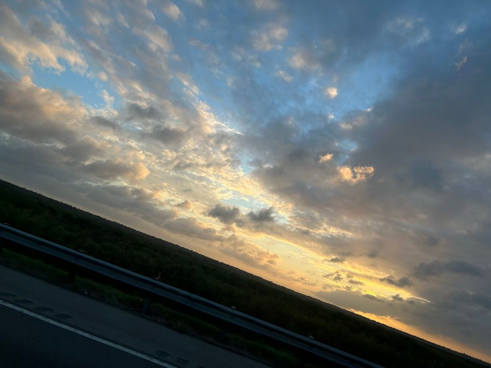 A golden sunset, surrounded by voluminous clouds. Shades of grey, white and gold show the contours of the clouds. Dark and backlit, a watery mash stretches out from freeway from which the photo was taken. 