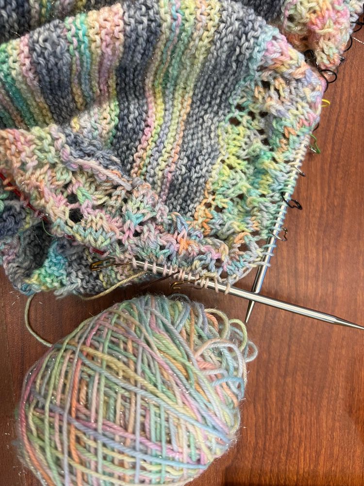 A piece of knitting sits on a wooden table. The knitter is halfway through a row, you can see the needles sticking out the top and stitch markers all down the side. The knitting is striped - alternating gray and rainbow sparkly vomit