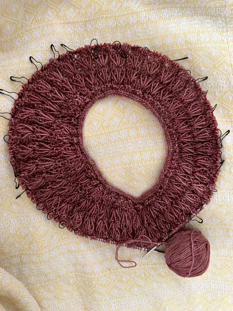 The yoke of a shirt lays on a light yellow blanket. It’s still small. The deep pink stitches are arranged like a sunburst around the circular needles.