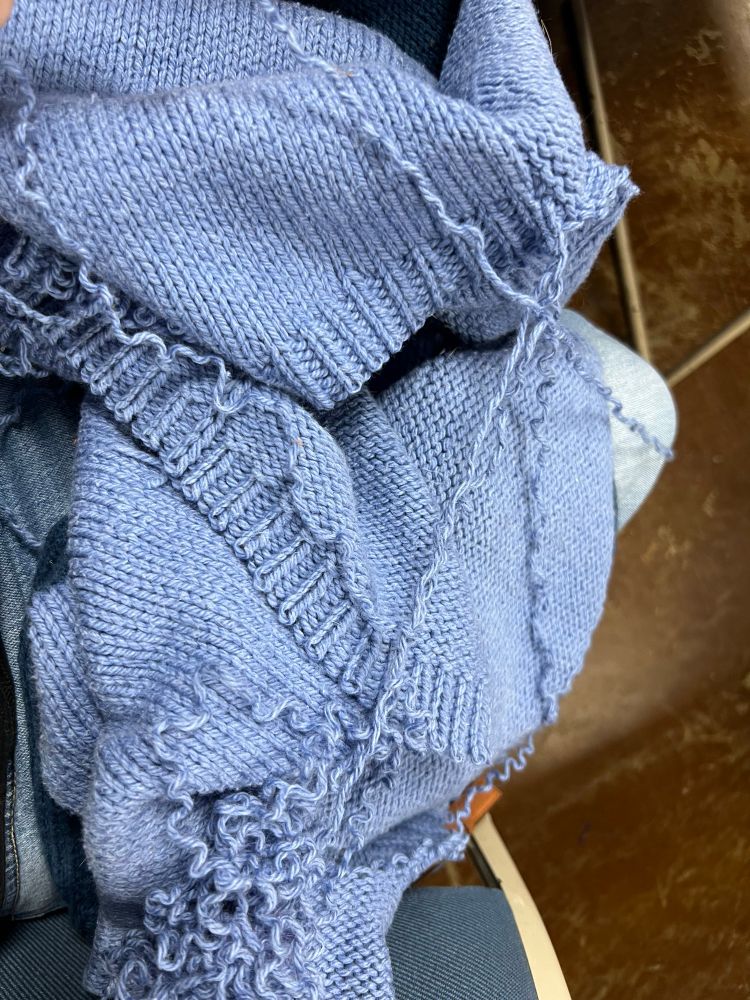 A blue sweater lays bunched up in a woman’s lap. There’s yarn hanging off one end