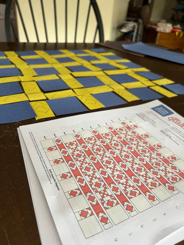 A quilt assembly diagram sits in the foreground, a sea of pink and white. Behind it, just out of focus, is a mess of blue and yellow squares and rectangles.