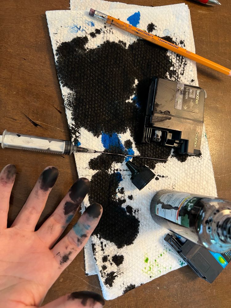 What a mess. A bottle of black ink and a long syringe lay on top of a paper towel, which is on a brown table. The paper towel is drenched in color - mostly large black splotches but some smaller blue and yellow ones as well. A white hand is palm up in the bottom corner. The fingers are also splotched.