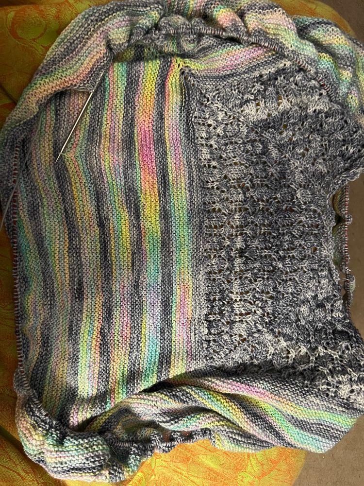 A piece of knitting is trying to lay flat but is gathered at the edges. The left portion is a lace pattern in mottled gray. On the right are a series of vertical stripes - alternating gray and rainbow sparkle vomit.