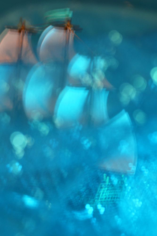 A bubbly, splashy ocean scene of blues and greens. Partially obscured by waves and bubbles, a little ship is in full sail with a jaunty green pennant fluttering atop a mast. This photo is a miniature ship surrounded by blue glitter tulle, with some ICM to smooth out the sails. 