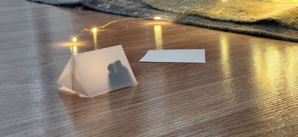 A little paper tent which may contain a silhouette of a cuddly Ed and Stede sits on a floor The last bulb on a of a string of fairy lights is experimentally illuminating the inside of the tent, the rest of the string lies haphazardly on the floor and extends out of frame.