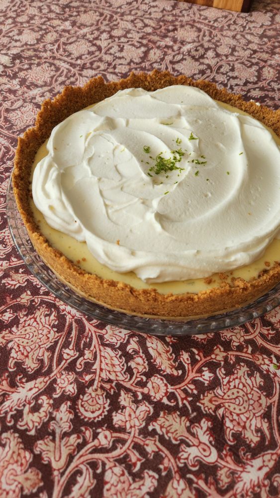 A key lime pie with a tall golden brown graham cracker crust, whipped cream, and golden filling looks appetizing on a table. 