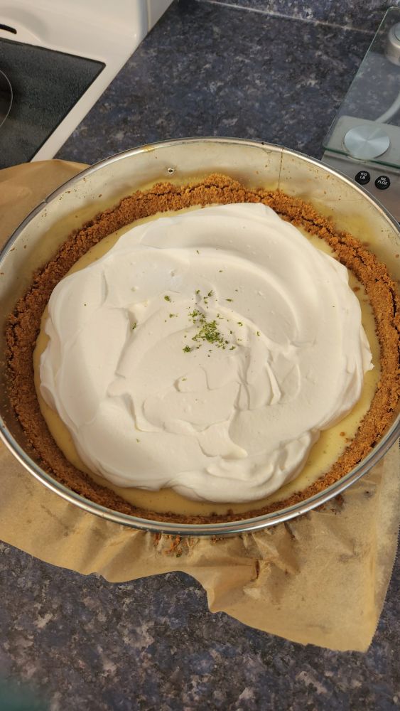 A key lime pie still in a Springform pan, with creamy white whipped cream and some sprinkles of lime zest on top. The crust is golden brown and the filling, which appears outside the edges of the whipped cream, is pale yellow.