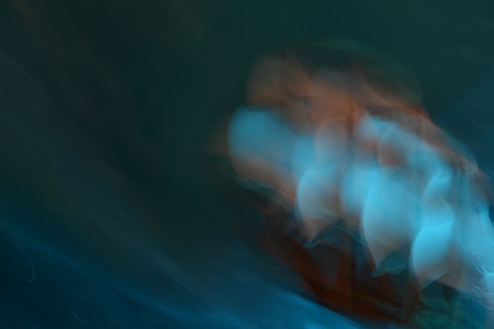 A murky, dark background resolves into flowing water under a wooden ship with billowing sails. Intentional camera movement has been used to create ghostly echoes of the sails, as though they are wrapped in gauze. The mood is dark, and the ship seems to be rushing urgently somewhere, even as it is dissolving. This is a photo of a small miniature ship and some fabric, with ICM to create the billowing sail effect. 