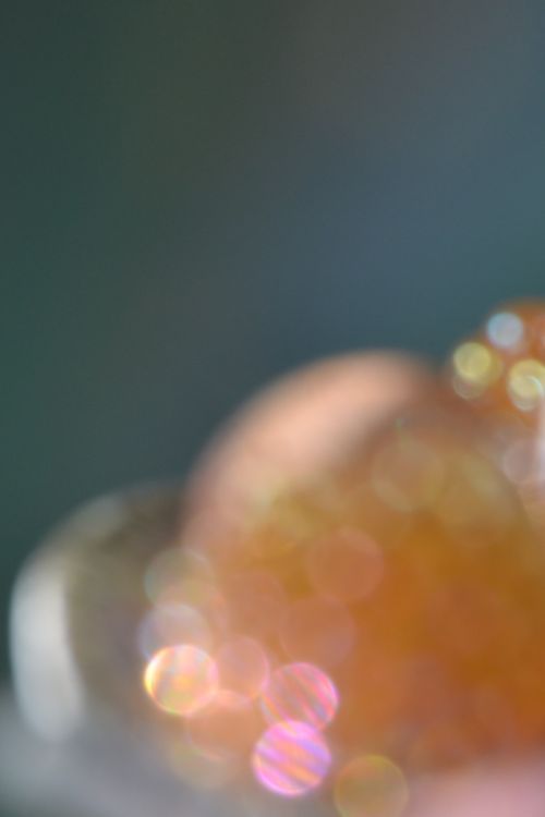 Against a slate blue background with hints of green and peach, a circular pale peach object is surrounded by a ring of grey that echoes the shape of the circle. Iridescent pink, yellow, orange and gold broken cluster around the circular shape, nearly obscuring it. Some of the largest pink bokeh in the foreground contains stripes of pink, orange, and blue. This photo was taken by placing costume jewellery in a white china dish of sand and photographing it very close up, so it's impossible to tell what the original objects are. The dish, peach circle, and bokeh create a pleasant rhythm of circles that repeats three times, running diagonally from the frame's lower left to the mid-right. The rest of the image is the slate background, creating a peaceful contrast with the bright colors below. The muted pinks and oranges contrast with the darker background, and the effect is peaceful and soothing (to me). 