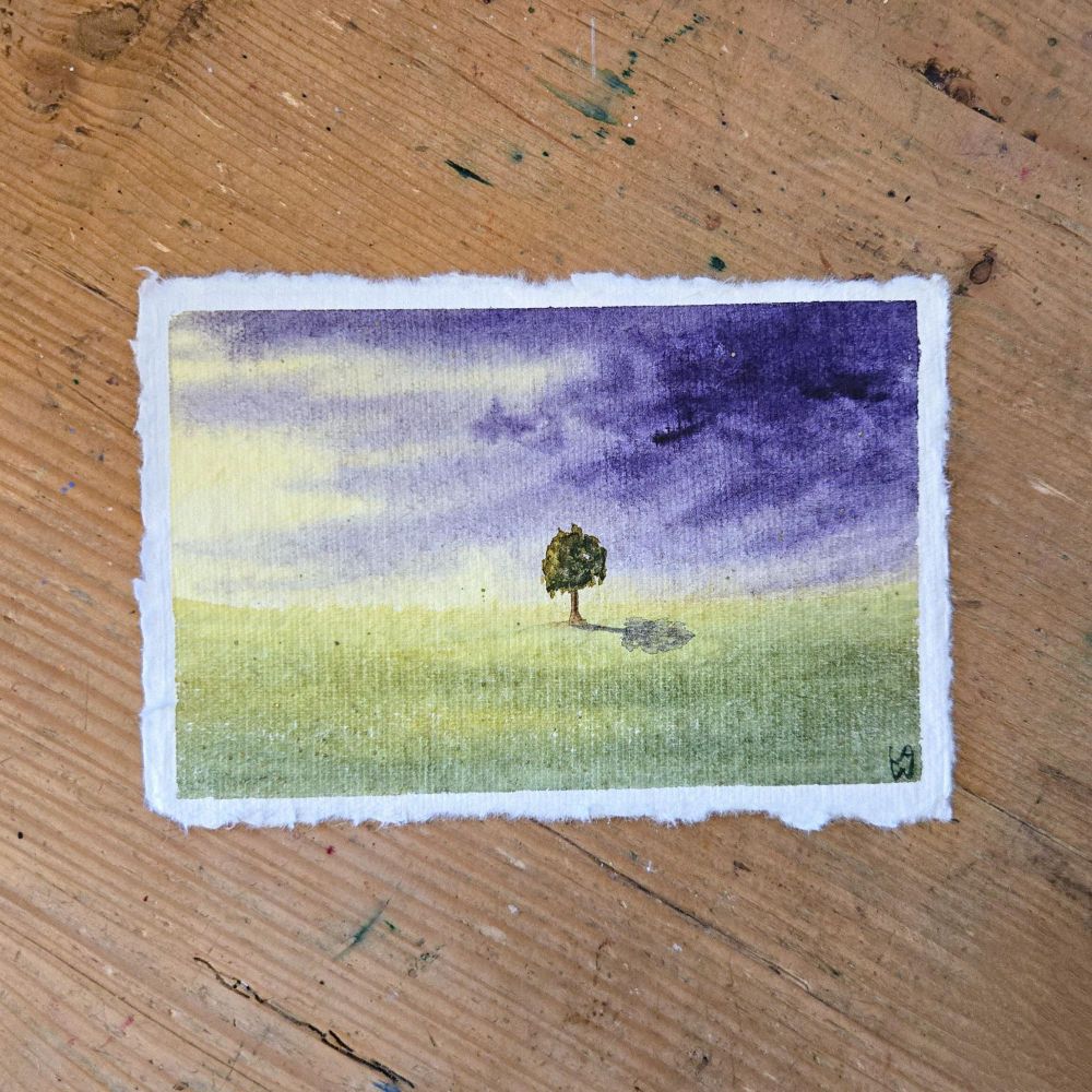 A watercolor painting on highly textured handmade paper. A single tree stands alone in the distance in the middle of a green field. On the right, dark purple storm clouds loom, with lighter purple clouds trailing to the left. On the left side, sunshine is beginning to peek out. 