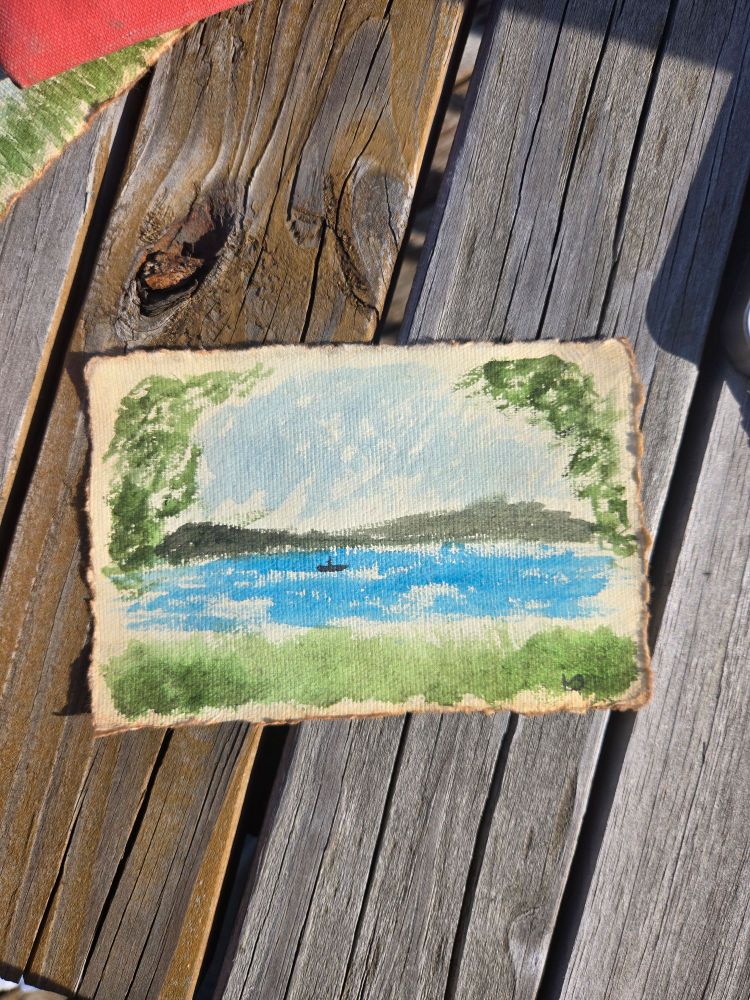 A watercolor painting sitting on a wooden table in the sunlight. The painting is done in a loose style and depicts a turquoise lake with a tiny rowboat in the center, some dark green hills in the background, and grass in the foreground. The top corners are framed with leaves from trees hanging overhead. 