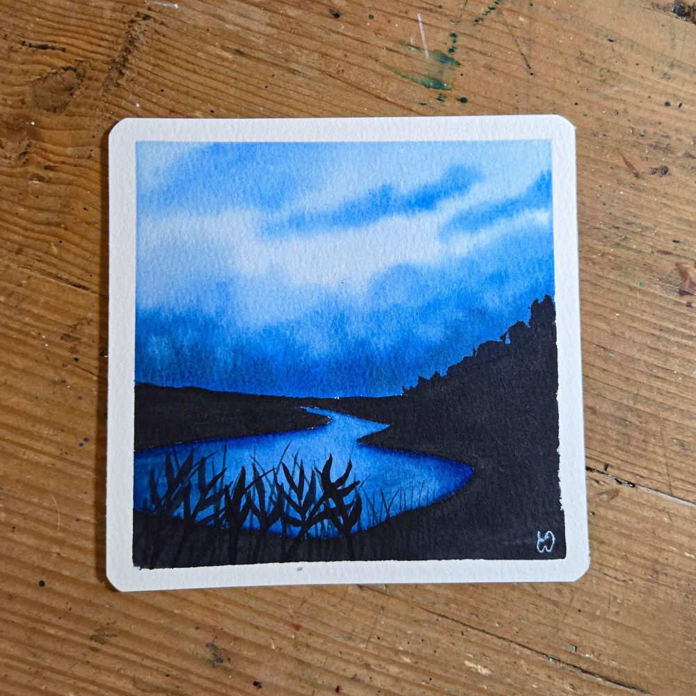 A moody watercolor landscape using phthalo blue and neutral tint. The blue sky is dark with clouds that are reflected in the lake below. The lake is surrounded by a very dark landscape with a hill on the right side and plants and grass in the foreground. 