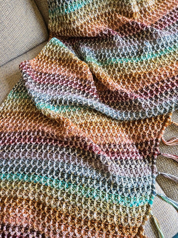 A close-up of the bottom of the shawl. The shawl is multicolored in a muted rainbow pattern. It is brioche knitted, with the lighter colors forming an elongated honeycomb pattern.