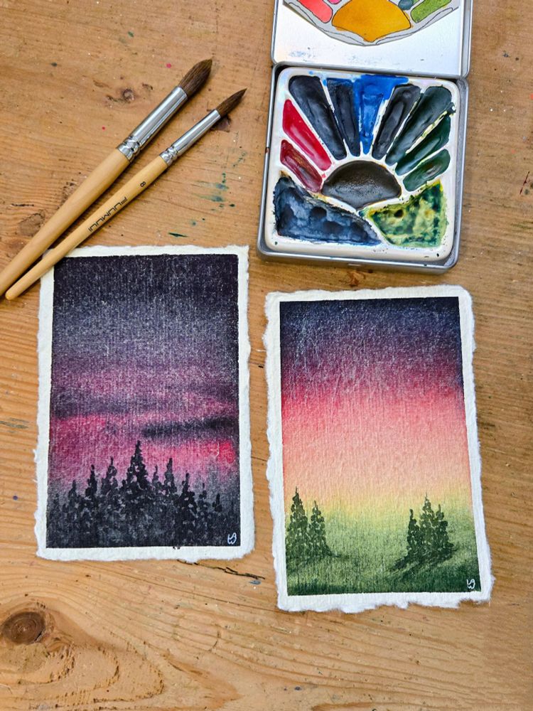 Two small watercolor paintings sit on a wooden desk, with a watercolor palette and two paint brushes. 

The painting on the left depicts a nighttime landscape, with an indigo sky and clouds, and a hint of magenta low on the horizon. In the foreground are pine trees silhouetted against the sky. 

The painting on the right depicts the last moments of sunset, with bright yellow on the horizon, followed by orange, pink, purple, and finally indigo as night descends. In the foreground are green hills with two distant groups of dark green pine trees.