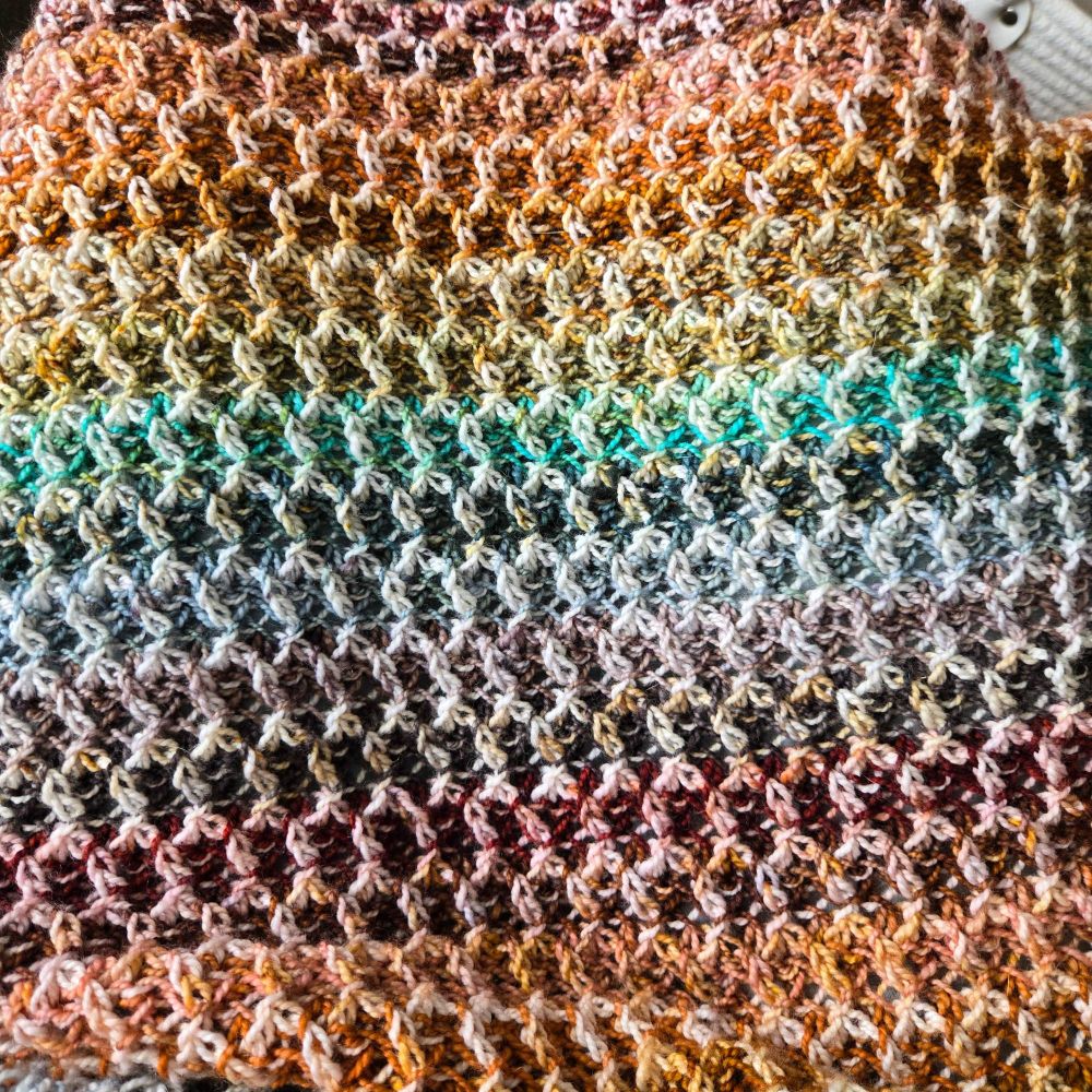 A brioche knitted shawl laying flat. It is multicolored with a kind of muted rainbow stripe pattern, and the brioche stitch pattern creates a honeycomb texture.