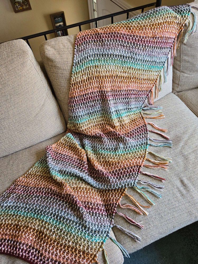 A long shawl with fringe on one long side resting on a couch. The shawl is multicolored in a muted rainbow pattern. It is brioche knitted, with the lighter colors forming an elongated honeycomb pattern.