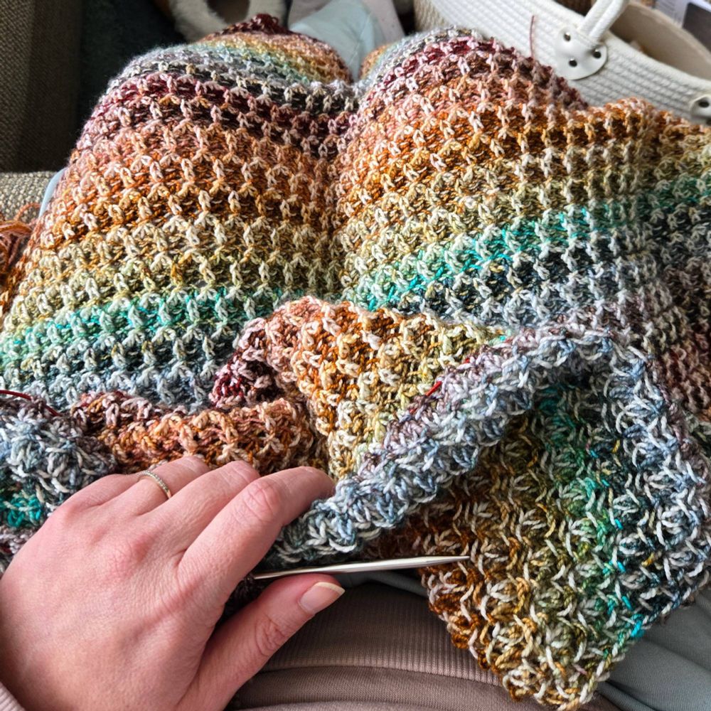A multicolored brioche knitted shawl sitting in a woman's lap. Her left hand rests on top holding a knitting needle. 