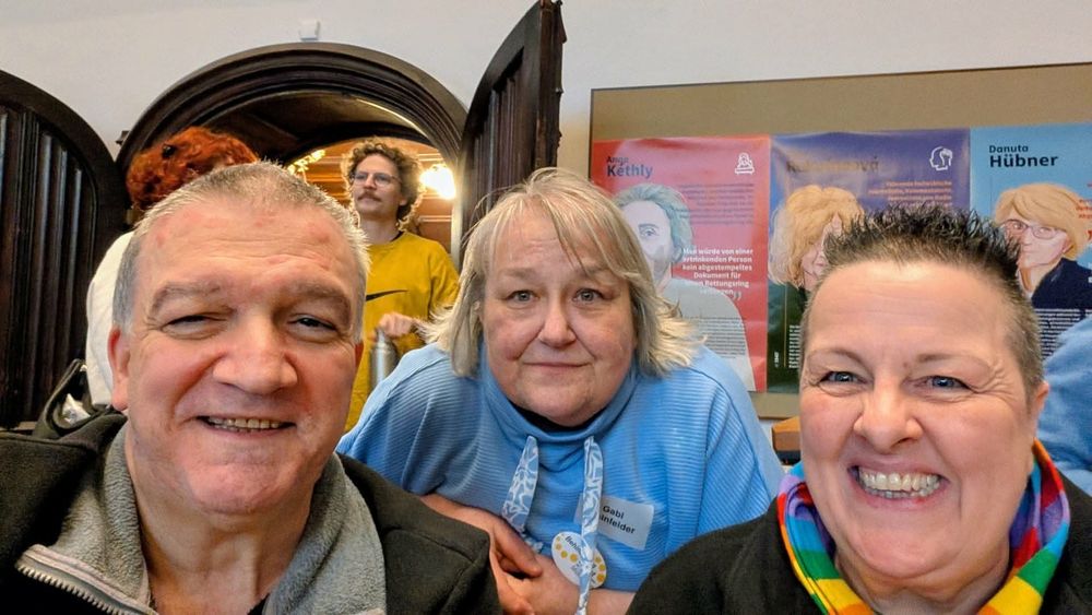 
Drei Personen sitzen an einem Tisch in einem Raum mit Holzdecke und bunten Plakaten an der Wand.