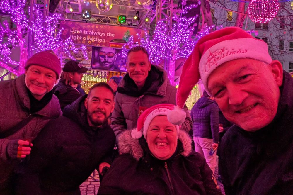 
Gruppe von Menschen bei Nacht auf einem Weihnachtsmarkt mit pink beleuchteten Bäumen und Weihnachtsmütze auf zwei Personen.