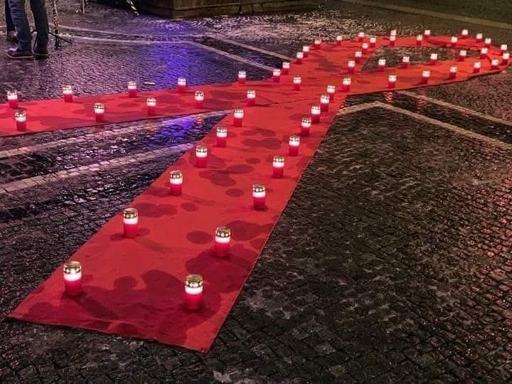 Roter Stoff in Form eines großen Schleifenbands auf Kopfsteinpflaster, darauf mehrere brennende Kerzen in Glasbehältern verteilt.