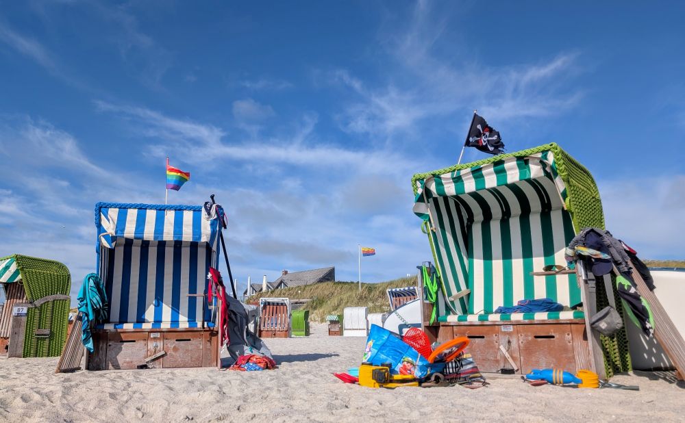 Auf dem Bild ist ein sonniger Strand zu sehen. Im Vordergrund stehen zwei typische norddeutsche Strandkörbe – der linke ist blau-weiß gestreift, der rechte grün-weiß. Beide sind mit bunten Handtüchern, Kleidung und Spielsachen dekoriert, was auf eine Familie oder Gruppe am Strand hinweist. Über dem linken Strandkorb weht eine Regenbogenflagge, über dem rechten eine Piratenflagge. Im Hintergrund sieht man weitere Strandkörbe, eine Düne mit Gras und ein Haus mit Schrägdach. Der Himmel ist blau mit einigen weißen Wolken. Die Szene wirkt lebendig und sommerlich.